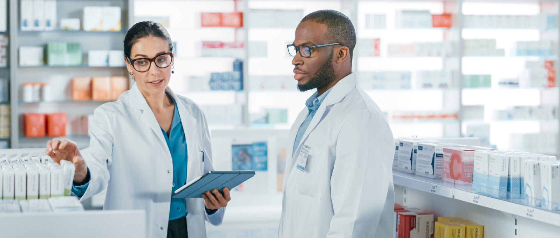 female pharmacist talking to a male pharmacist while getting a bottle of pills and holding a tablet
